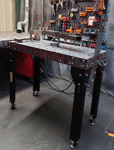 A heavy duty modular welding table with black powder-coated legs and a bare steel top, equipped with lockable castor wheels and various tools placed on top.